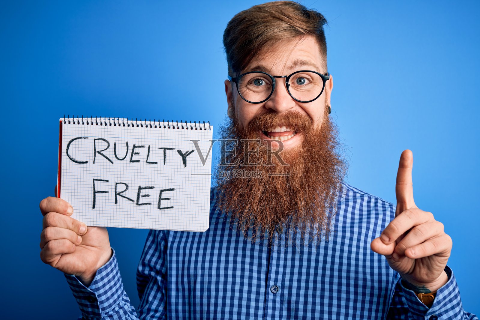 在蓝色背景前拿着纯素化妆品信息的红发爱尔兰男人，惊喜地指着手指照片摄影图片