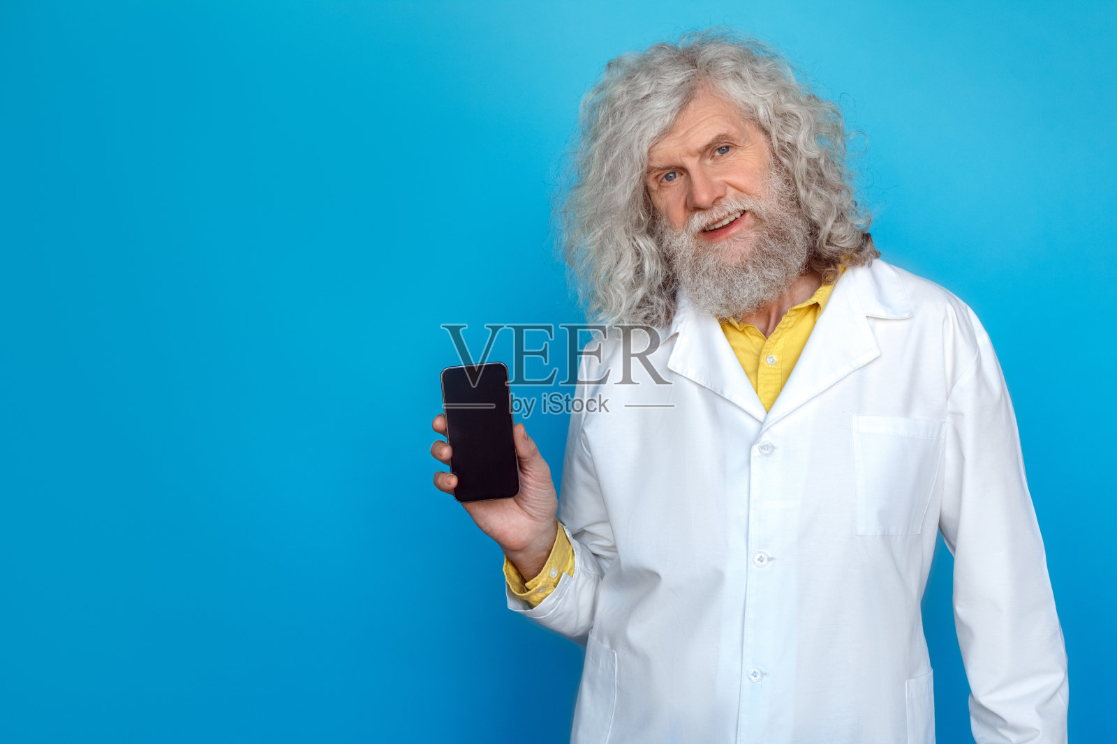 穿着医生袍的老人在蓝色背景上隔离，向相机展示智能手机，面带微笑照片摄影图片