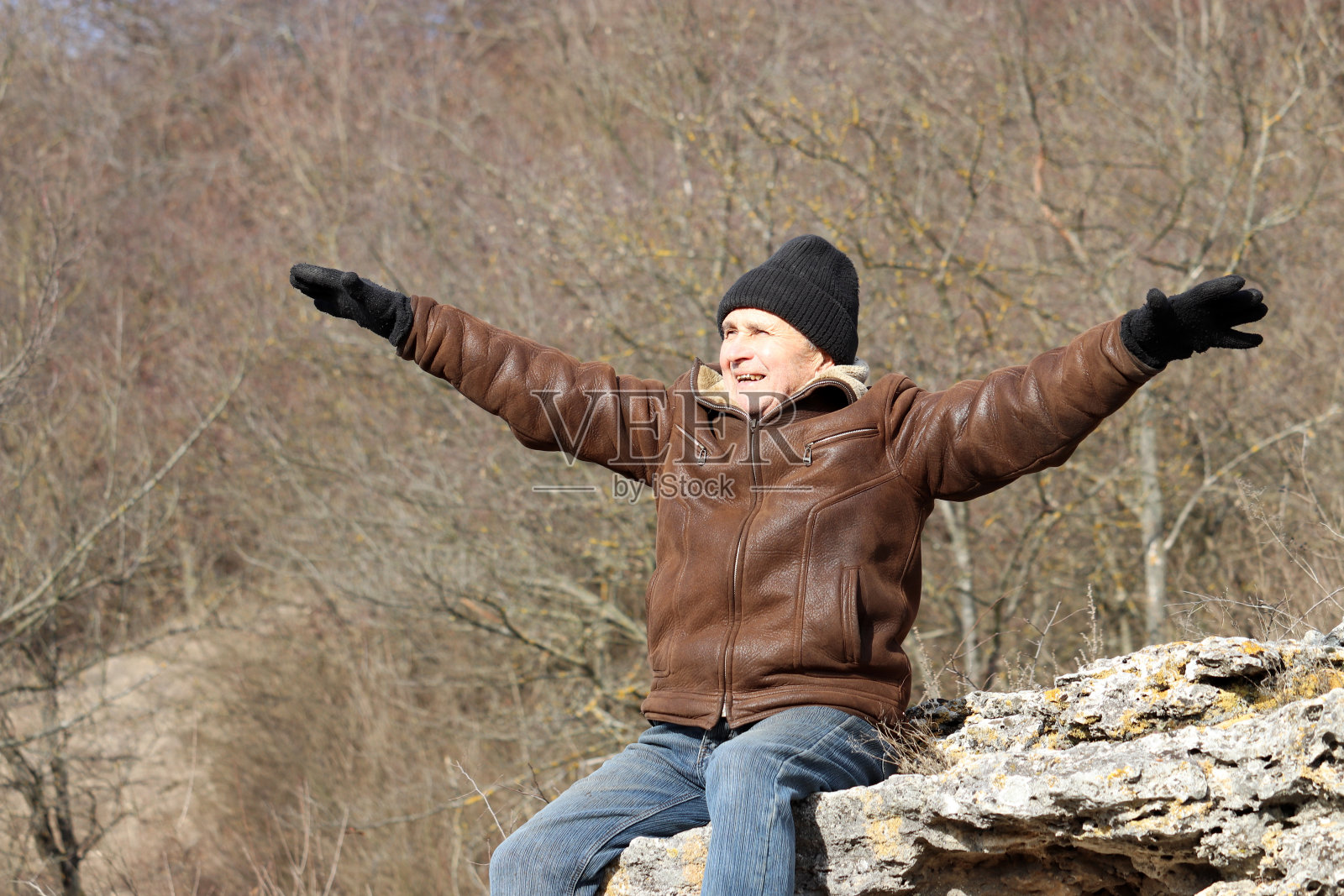 一位穿着牛仔裤和皮衣坐在森林岩石上的快乐老人举起了手照片摄影图片