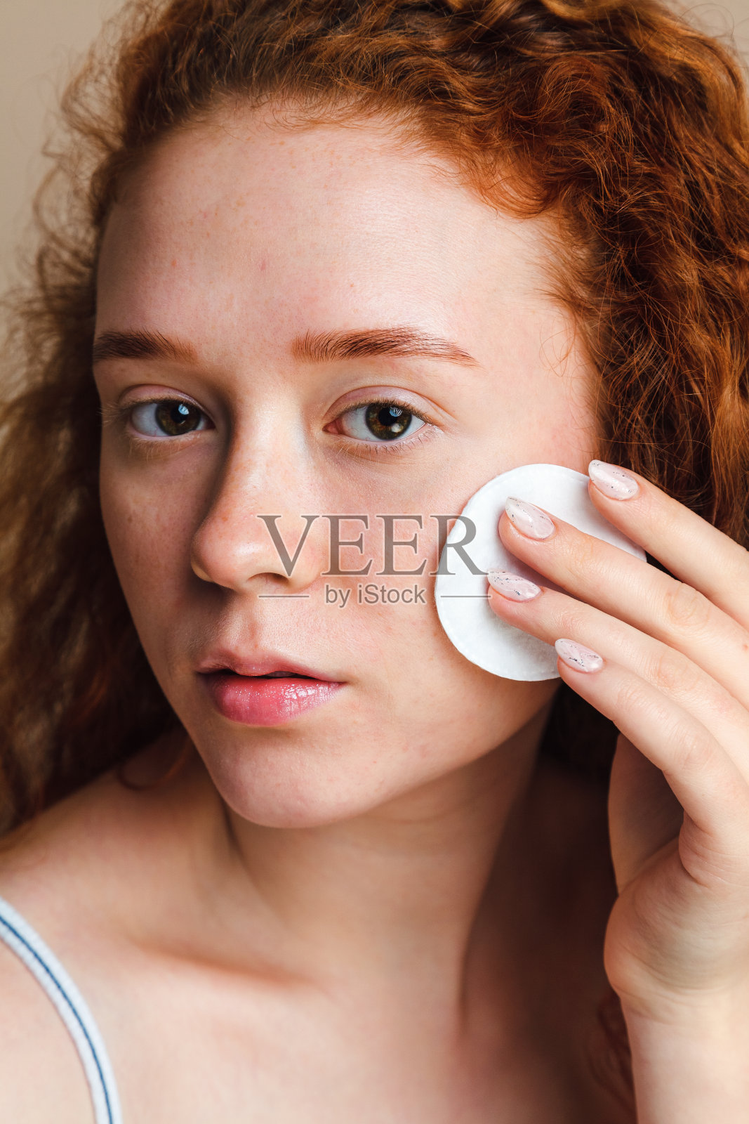 年轻护肤理念。迷人的少女与长长的红色鬈发摩擦她的脸与棉垫。不化妆，不修饰。自然美景。照片摄影图片