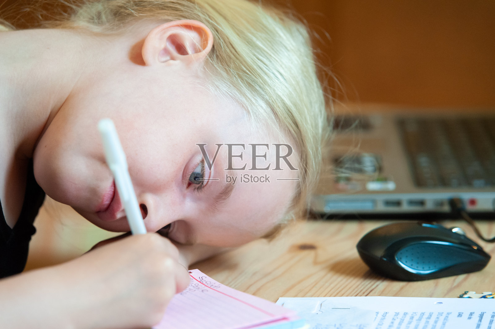 年轻的金发女孩低着头写着。笔记本电脑和鼠标在背景中失去焦点照片摄影图片