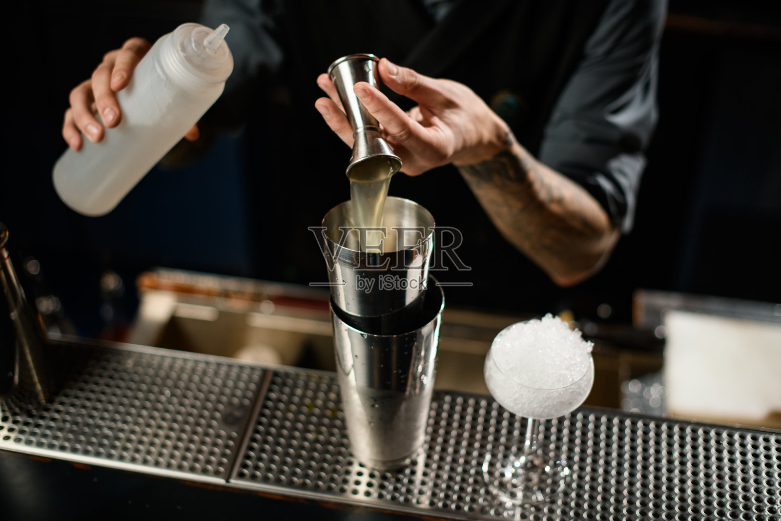 调酒师将酒精饮料从摇酒器倒到一个钢制摇酒器上，摇酒器上握着一个白色塑料瓶照片摄影图片
