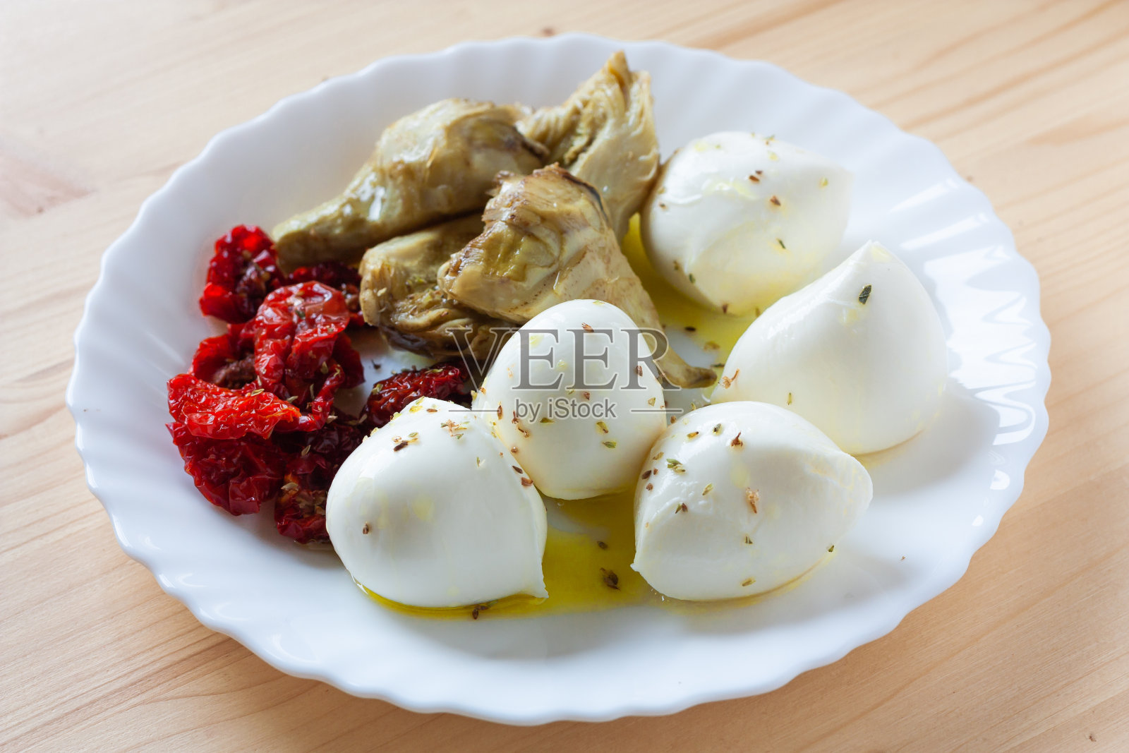 木桌上的白盘子上放着马苏里拉奶酪、干樱桃番茄和用橄榄油和牛至调味的洋蓟。照片摄影图片