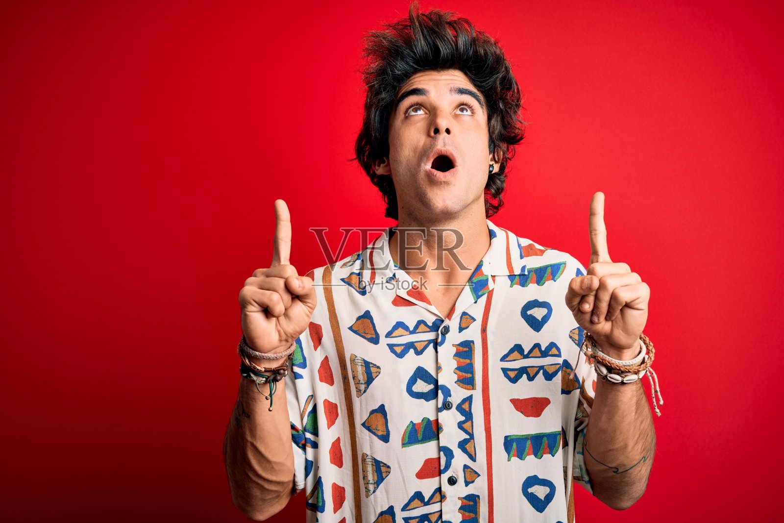年轻英俊的男人在假期穿着夏天的衬衫在孤立的红色背景惊讶和惊讶的看着和指向手指和举起的手臂。照片摄影图片
