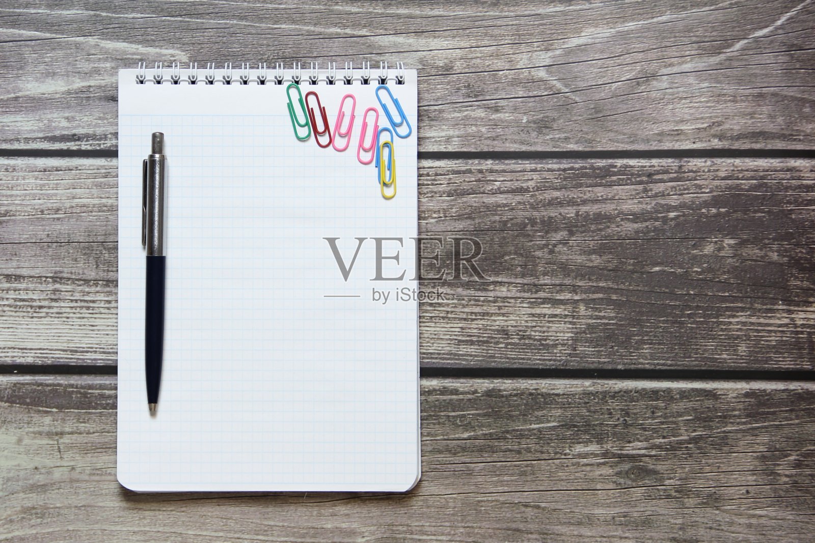 在木板的背景上放着一个记事本，上面有一张空白的白纸，还有圆珠笔和回形针照片摄影图片