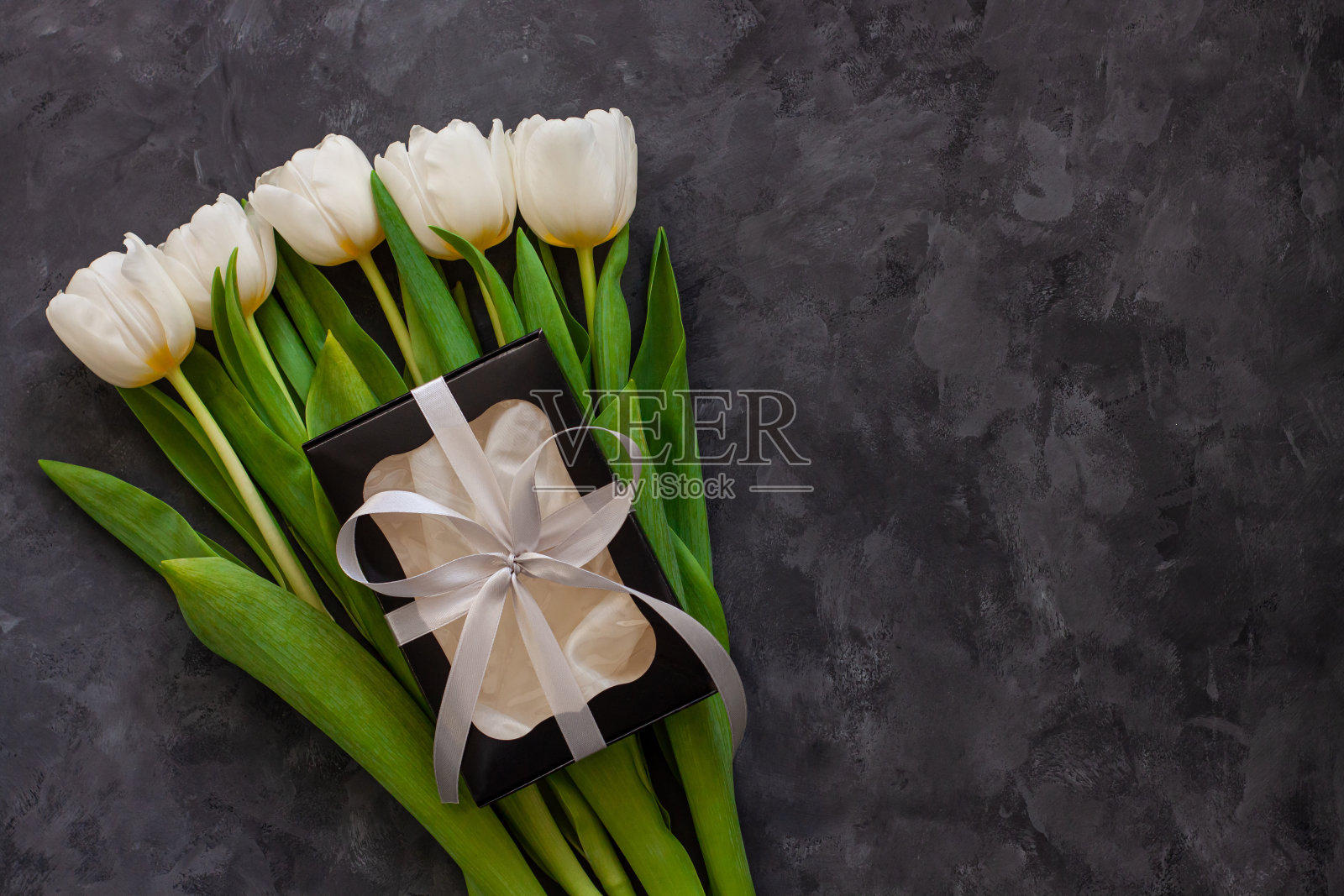 白色的郁金香花和黑色的礼盒在深色的背景上平躺着。三月八日妇女节快乐。照片摄影图片