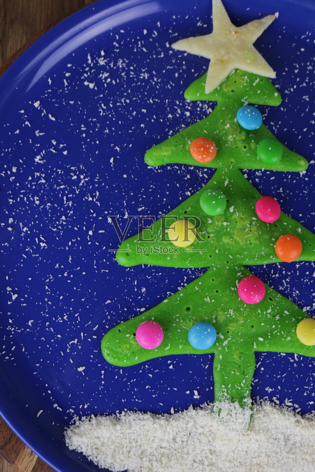 形象的绿色煎饼形状的圣诞树为圣诞节儿童的早餐，圣诞树煎饼装饰有彩虹色，糖衣巧克力甜，上面有圣诞星星，干燥的椰子冬天雪在蓝色盘子上。照片摄影图片