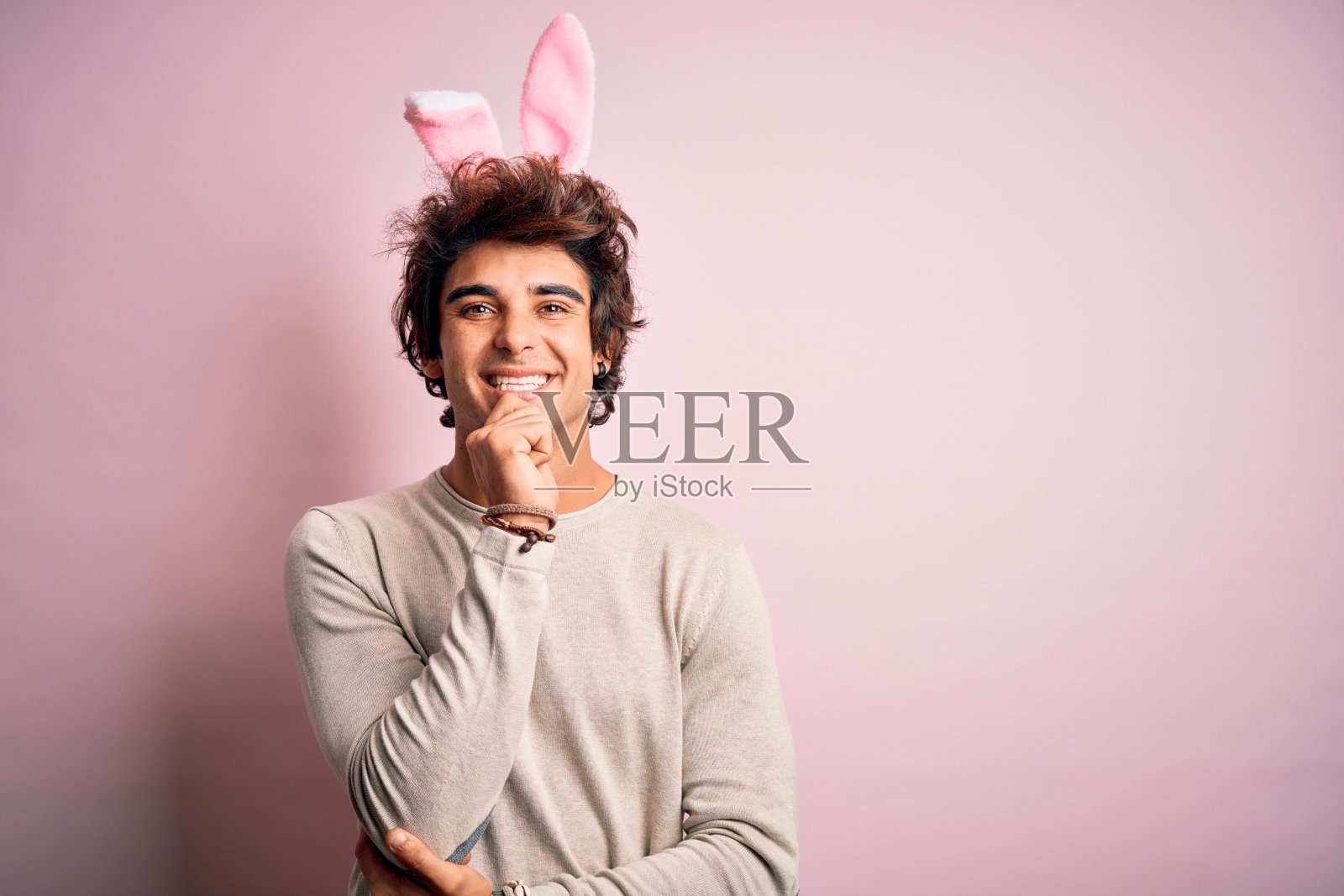 年轻英俊的男子抱着复活节兔子耳朵站在孤立的粉红色背景看起来自信在镜头与微笑交叉双臂和手高举在下巴。积极的思考。照片摄影图片