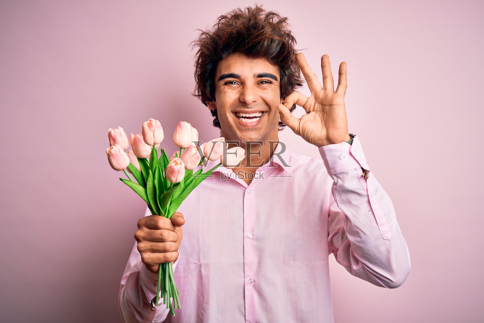 年轻英俊的男子拿着花站在孤立的粉红色背景做ok手势与手指，优秀的象征照片摄影图片