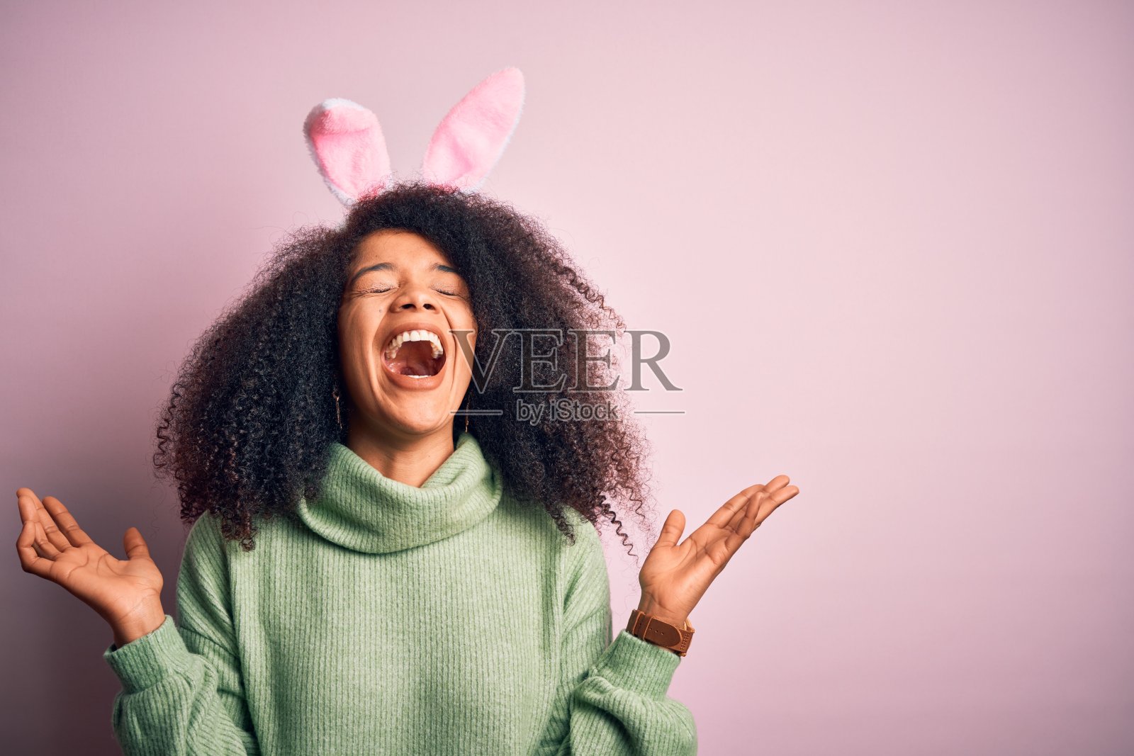 年轻的非洲裔美国女性，蓬松发型，粉色背景，戴着复活节兔子耳朵，举起双臂，闭着眼睛尖叫，庆祝成功。照片摄影图片