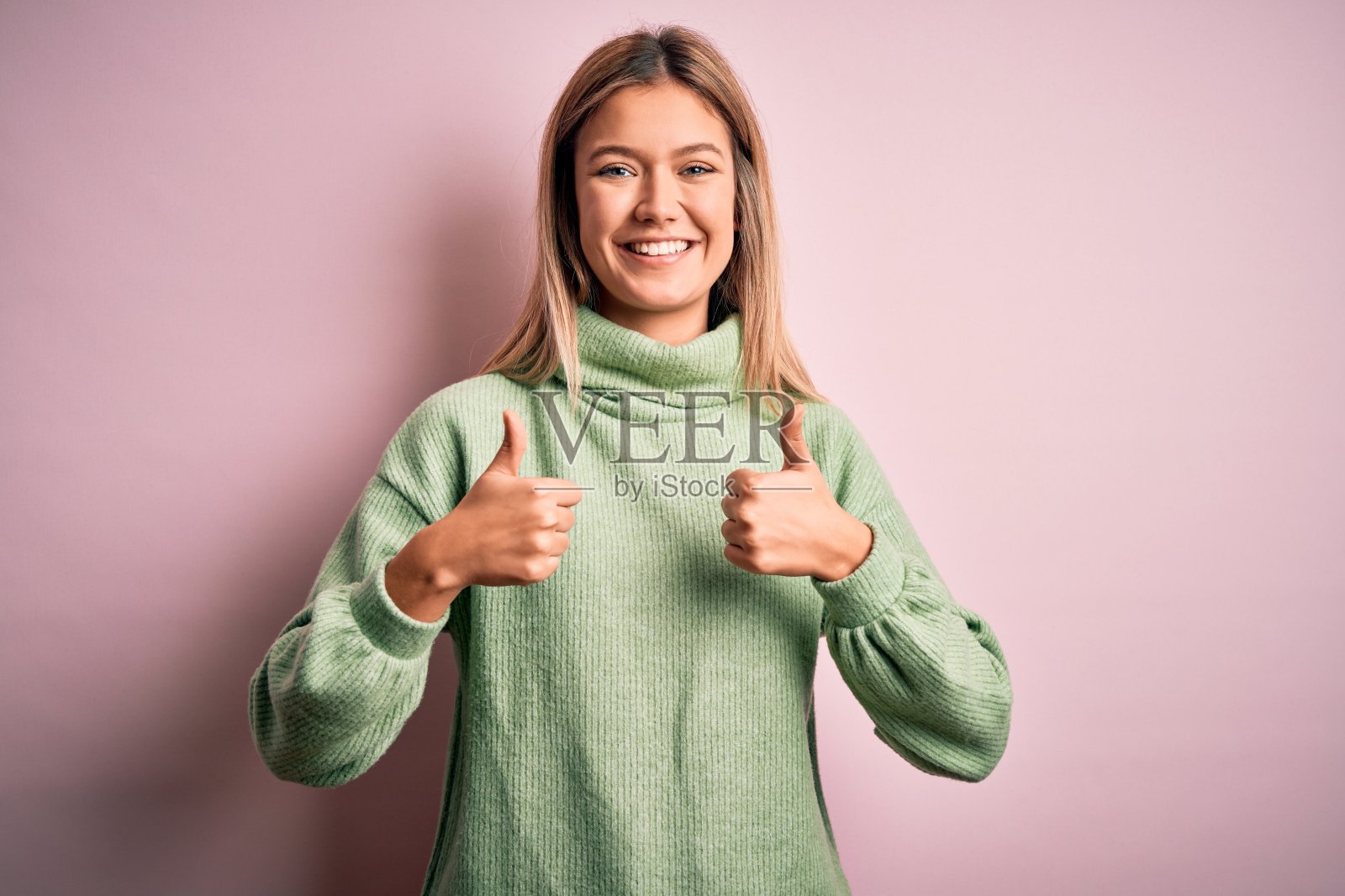 年轻美丽的金发女人穿着冬季羊毛衫在粉红色孤立的背景成功标志做积极的手势与手，拇指向上微笑和幸福。愉快的表情和胜利者的姿态。照片摄影图片