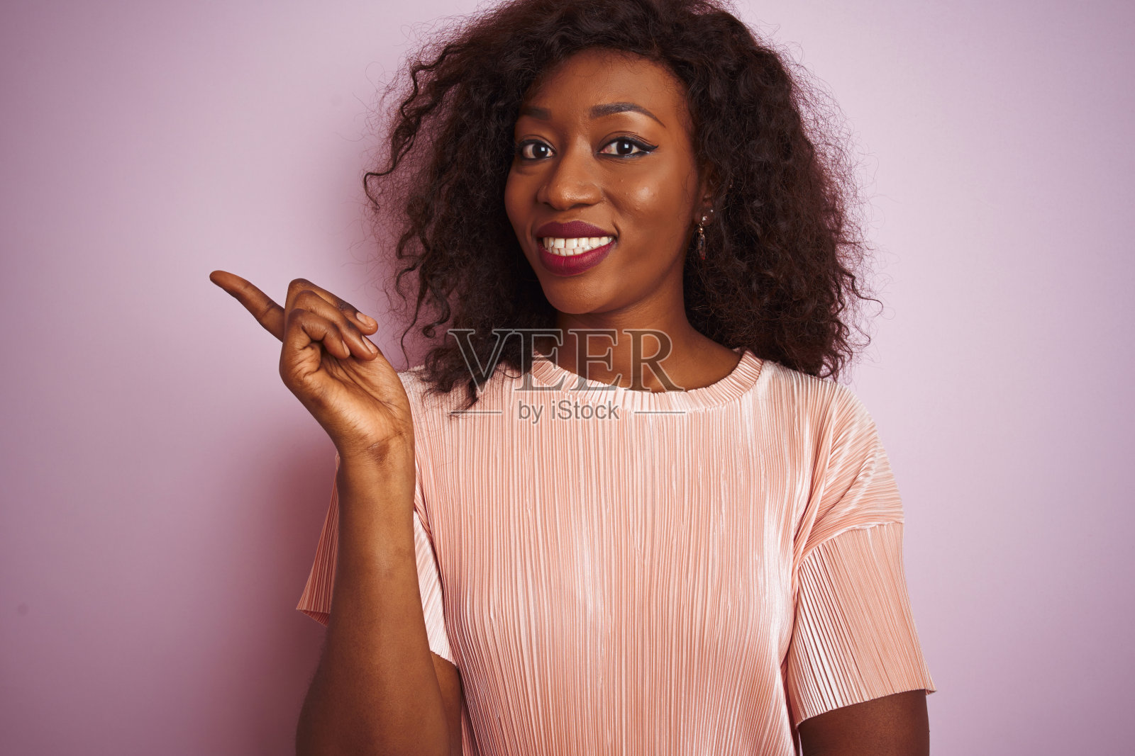 年轻的非洲裔美国妇女穿着t恤站在孤立的粉红色背景上，脸上有一个大大的微笑，用手和手指指着一边看着相机。照片摄影图片