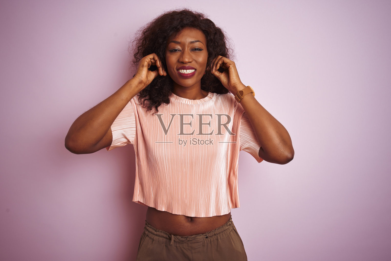 年轻的非洲裔美国妇女穿着t恤站在孤立的粉红色背景覆盖耳朵与手指与烦恼的表情大声的音乐噪音。聋的概念。照片摄影图片