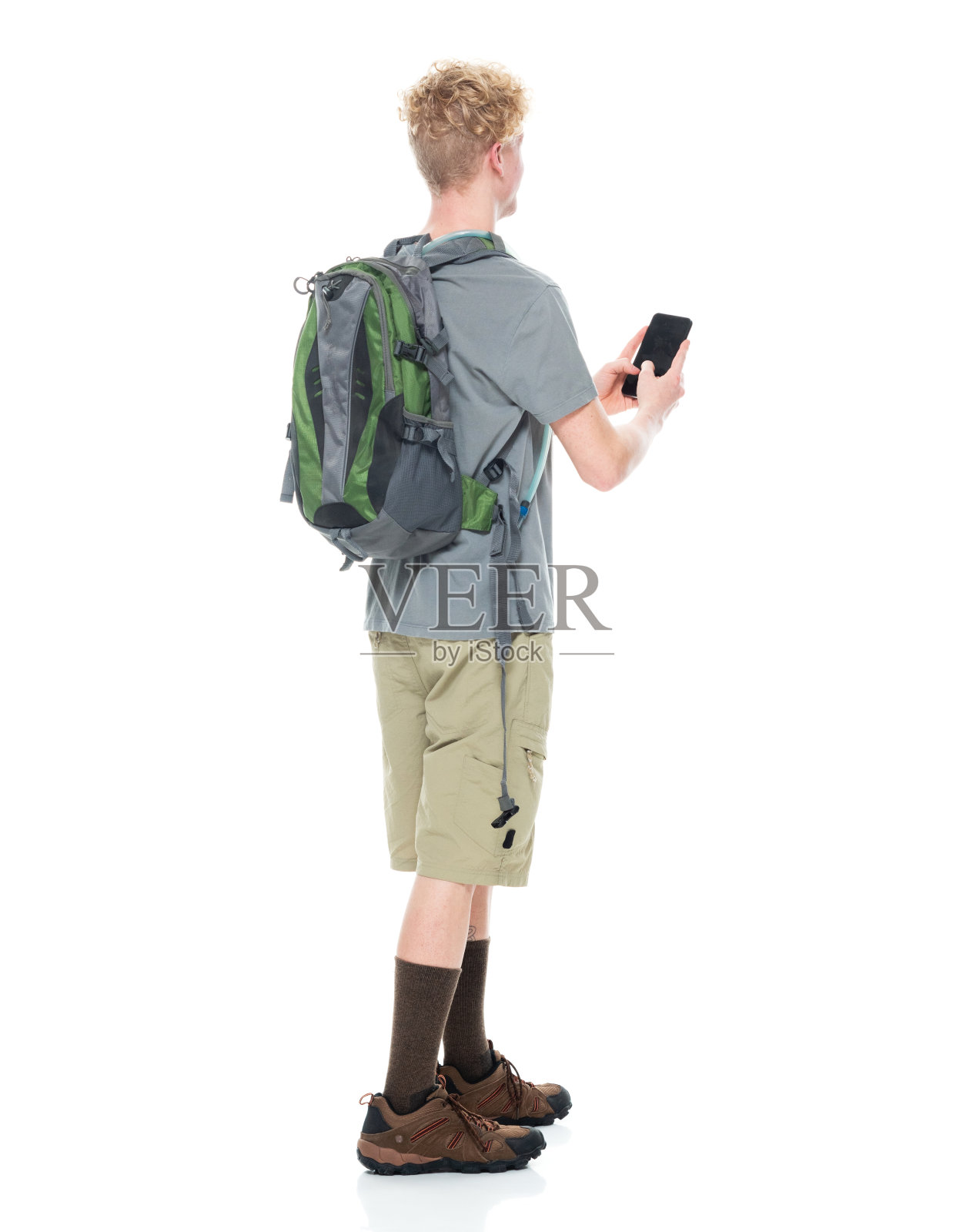 白人年轻男性背包客穿着登山靴，拿着包，用智能手机在白色背景前徒步旅行照片摄影图片