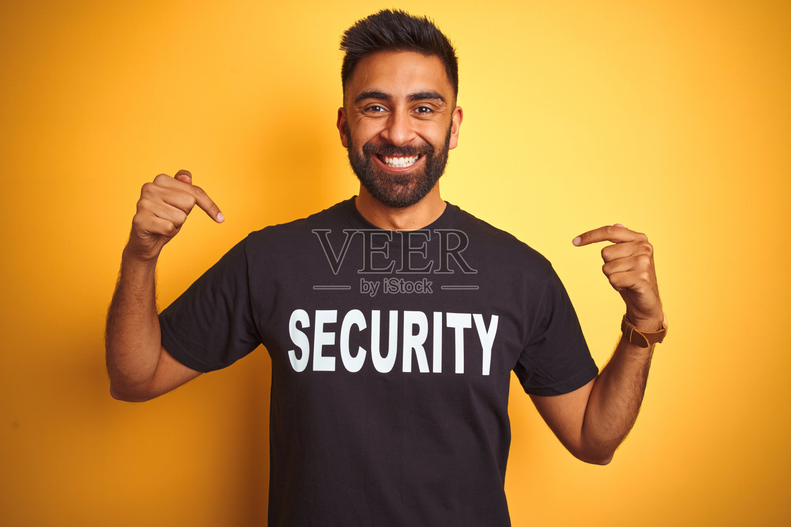 在黄色背景下身穿保安制服的阿拉伯裔印度混血保安，面带微笑，自信地用手指自豪地指向自己照片摄影图片