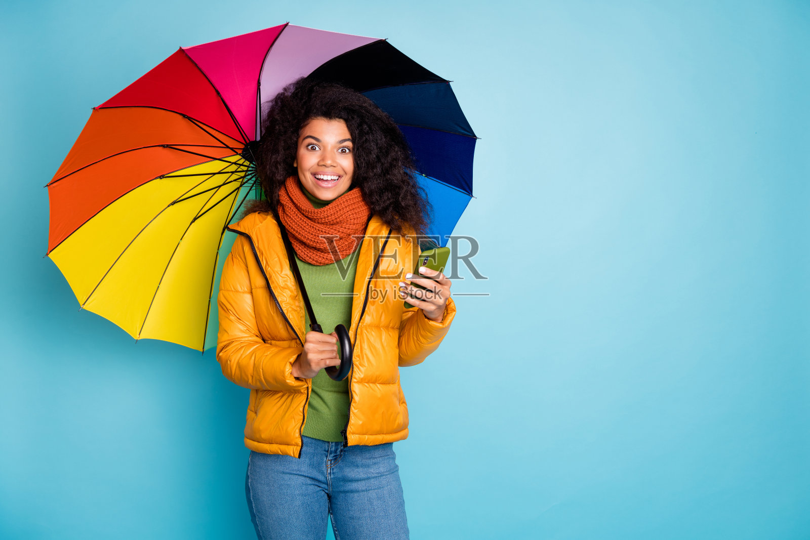 漂亮的深色皮肤女士的照片拿着五颜六色的伞电话现代的社交网络博主穿着时尚的大衣牛仔裤绿色套衫围巾孤立的蓝色背景照片摄影图片