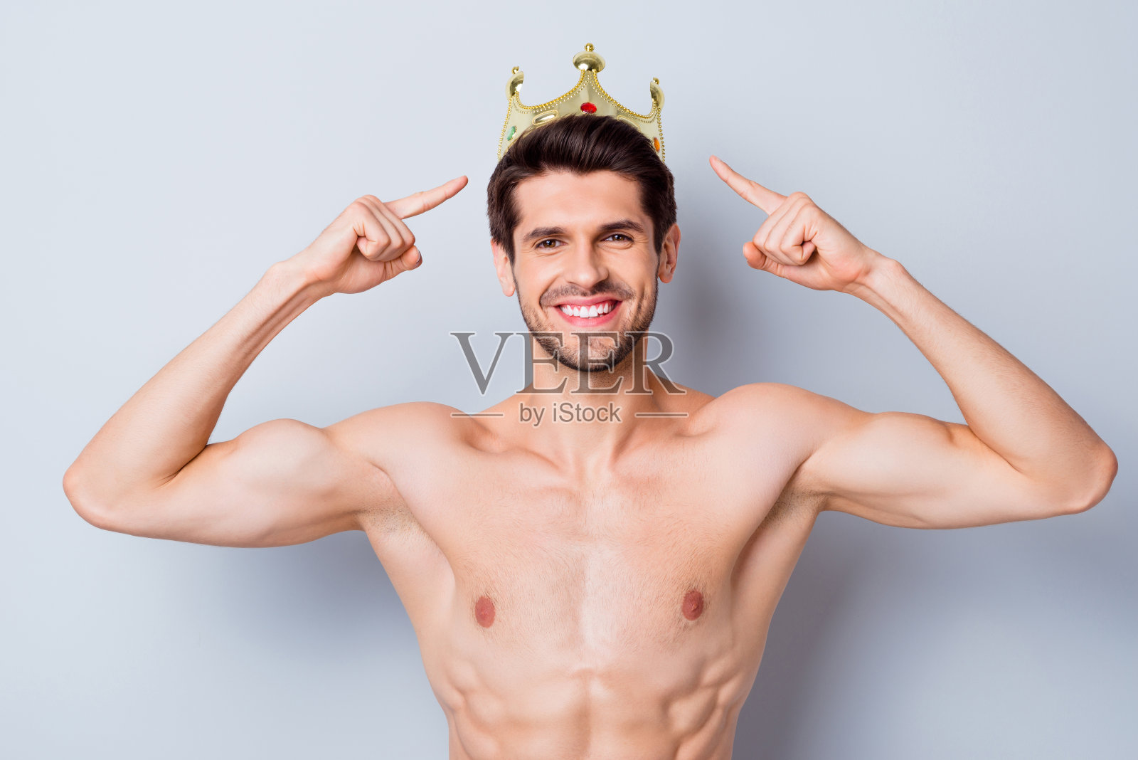 看，我是运动之王。英俊的男子强壮的家伙戴着金色的皇冠，他赢得举重运动比赛点食指停留在淋浴胸前浴孤立在灰色的背景照片摄影图片