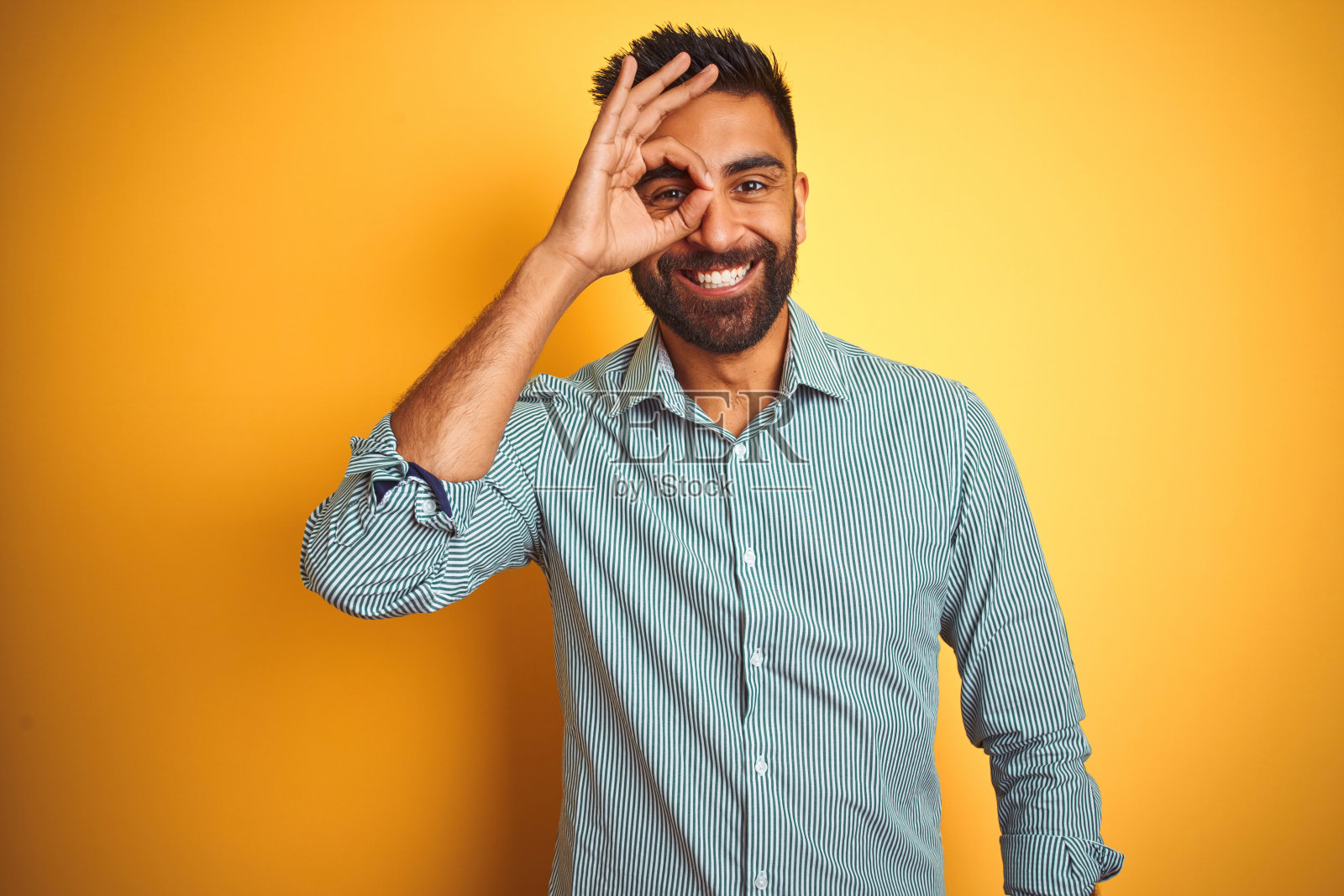在黄色背景前，身穿绿色条纹衬衫的年轻印度男子用手做出OK手势，面带微笑，眼睛透过手指看，面露喜色照片摄影图片