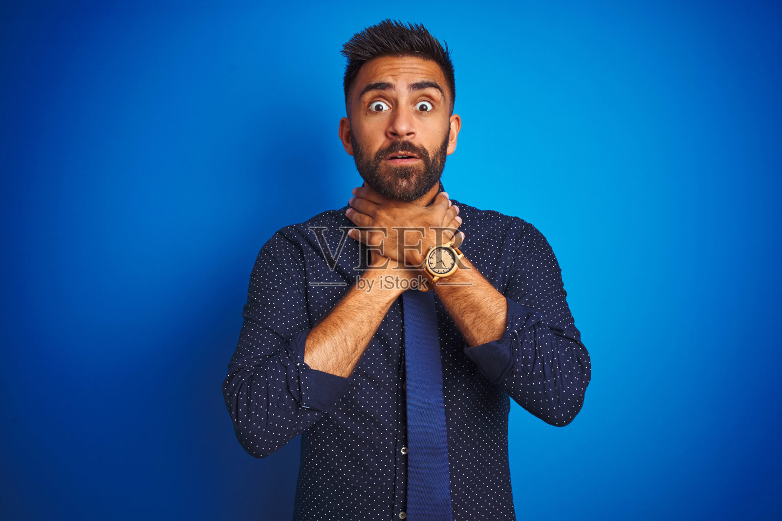 印度年轻商人穿着优雅的衬衫和领带站在孤立的蓝色背景下，因痛苦的扼杀而尖叫和窒息。健康问题。窒息和自杀概念照片摄影图片