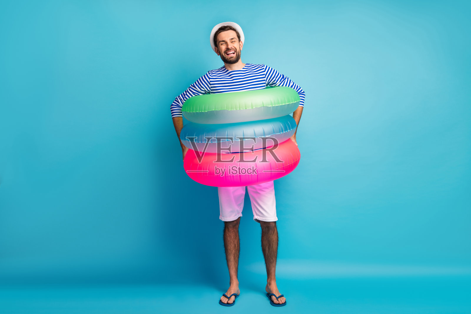 时髦的家伙游客的全长照片进入三个彩色的橡胶救生圈去游泳池海洋穿条纹水手衬衫帽子短裤flip flops孤立的蓝色背景照片摄影图片