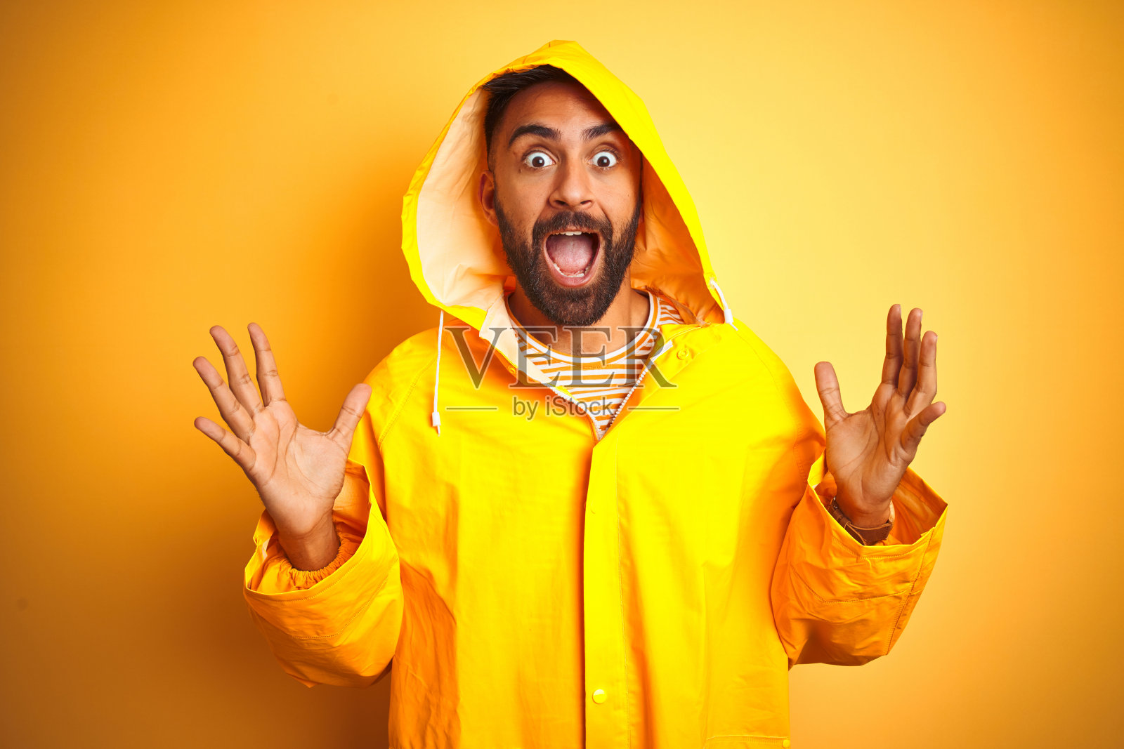 年轻的印度男人身穿带帽雨衣，站在黄色背景前，疯狂地庆祝成功，举起双臂，睁大眼睛，兴奋地尖叫，是成功者的概念照片摄影图片