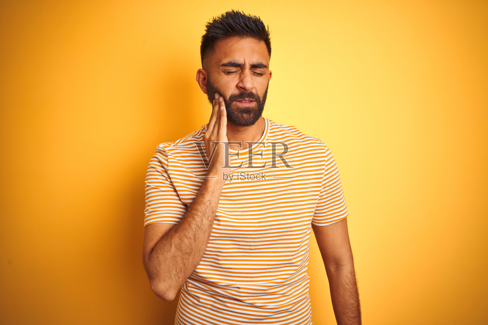 年轻印度男子，身穿T恤，在黄色背景前用手触摸嘴巴，表情痛苦，因为牙痛或牙齿疾病，牙医概念照片摄影图片