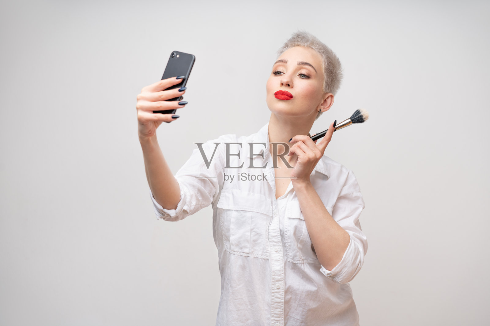 一位留着短发、化着妆的时髦女孩，手扶腰部，用手机自拍的影棚肖像照片摄影图片