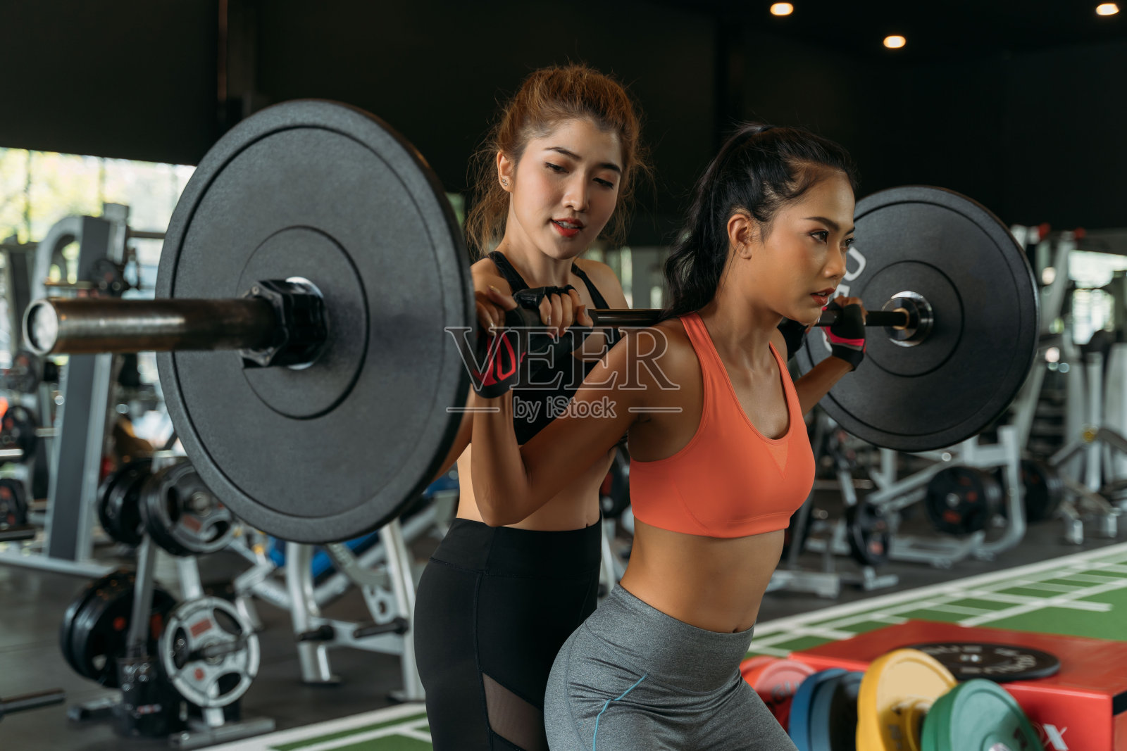 亚洲私人教练指导健美女性在健身房进行杠铃深蹲照片摄影图片