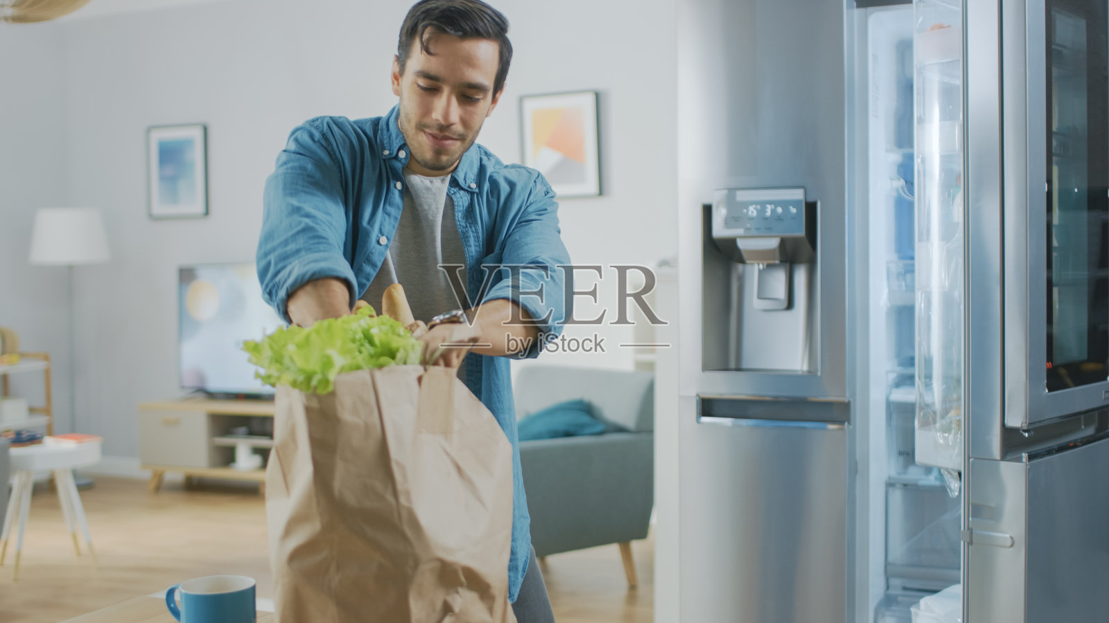 英俊迷人的年轻人把食品杂货带到厨房。他买了新鲜的蔬菜沙拉。现代冰箱是背景。房间有明亮的现代内饰。照片摄影图片