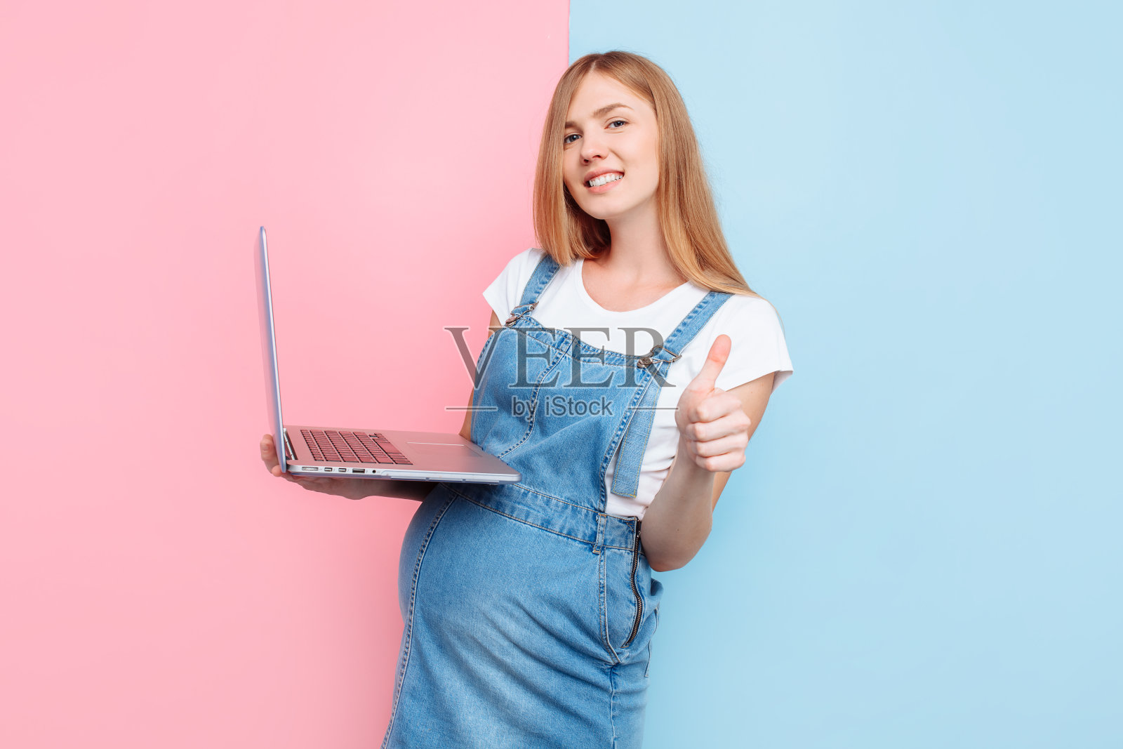一个快乐的孕妇站在一个孤立的粉色和蓝色背景上使用笔记本电脑的照片照片摄影图片