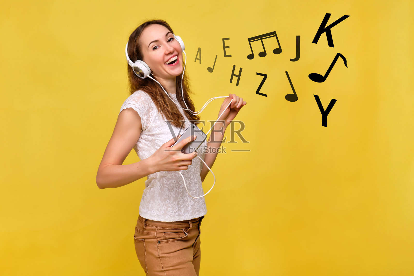 穿着夏装的女孩站在黄色背景前，戴着白色耳机听音乐，拿着智能手机唱歌。抽象的音符和字母从嘴里冒出来照片摄影图片