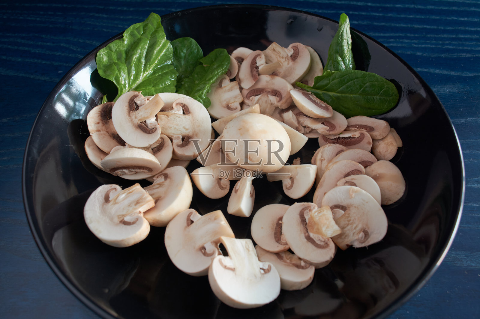 整个蘑菇和切片，放在黑色的盘子里，配上绿色的菠菜叶，在蓝色的背景上照片摄影图片