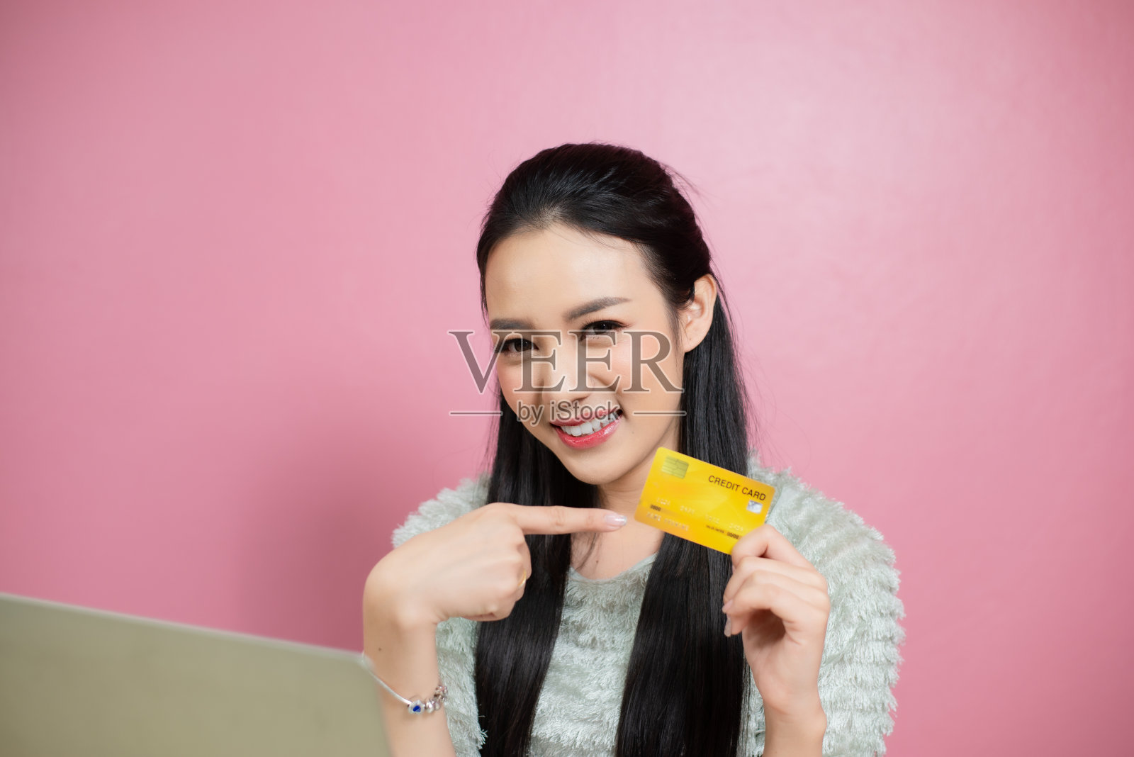 美丽亚洲笑脸女子指着信用卡粉红色的背景。照片摄影图片