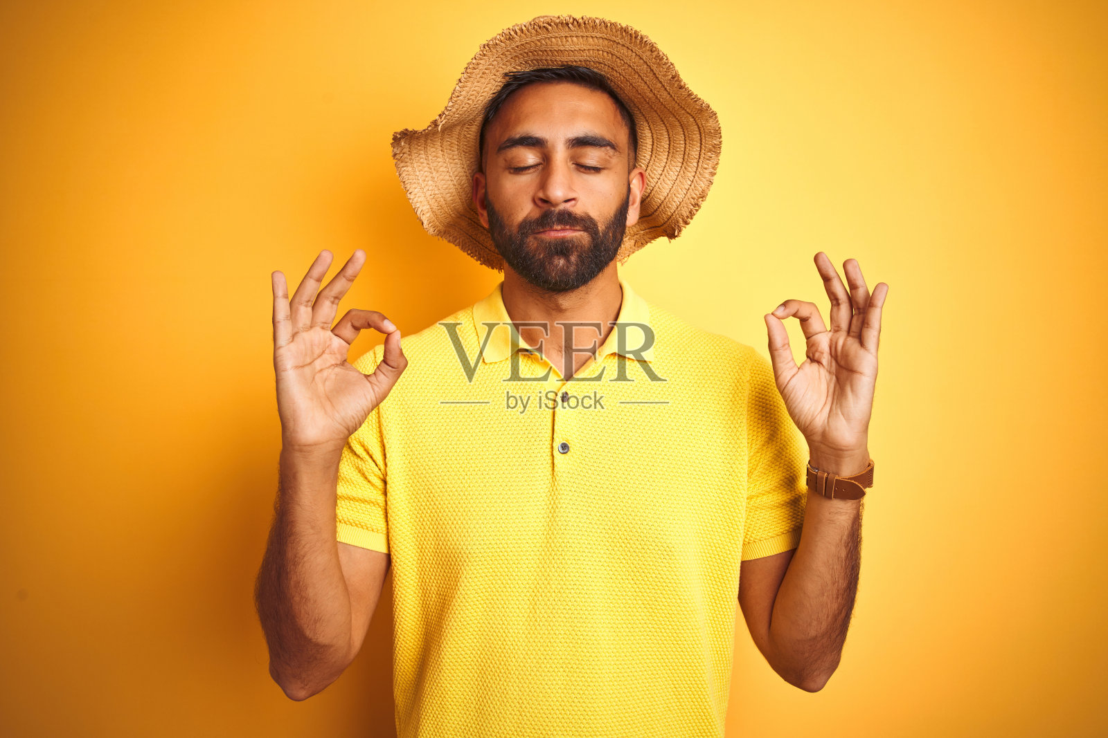 印度年轻男子度假，头戴夏帽，在黄色背景前放松微笑，闭眼冥想，手指做出瑜伽姿势照片摄影图片