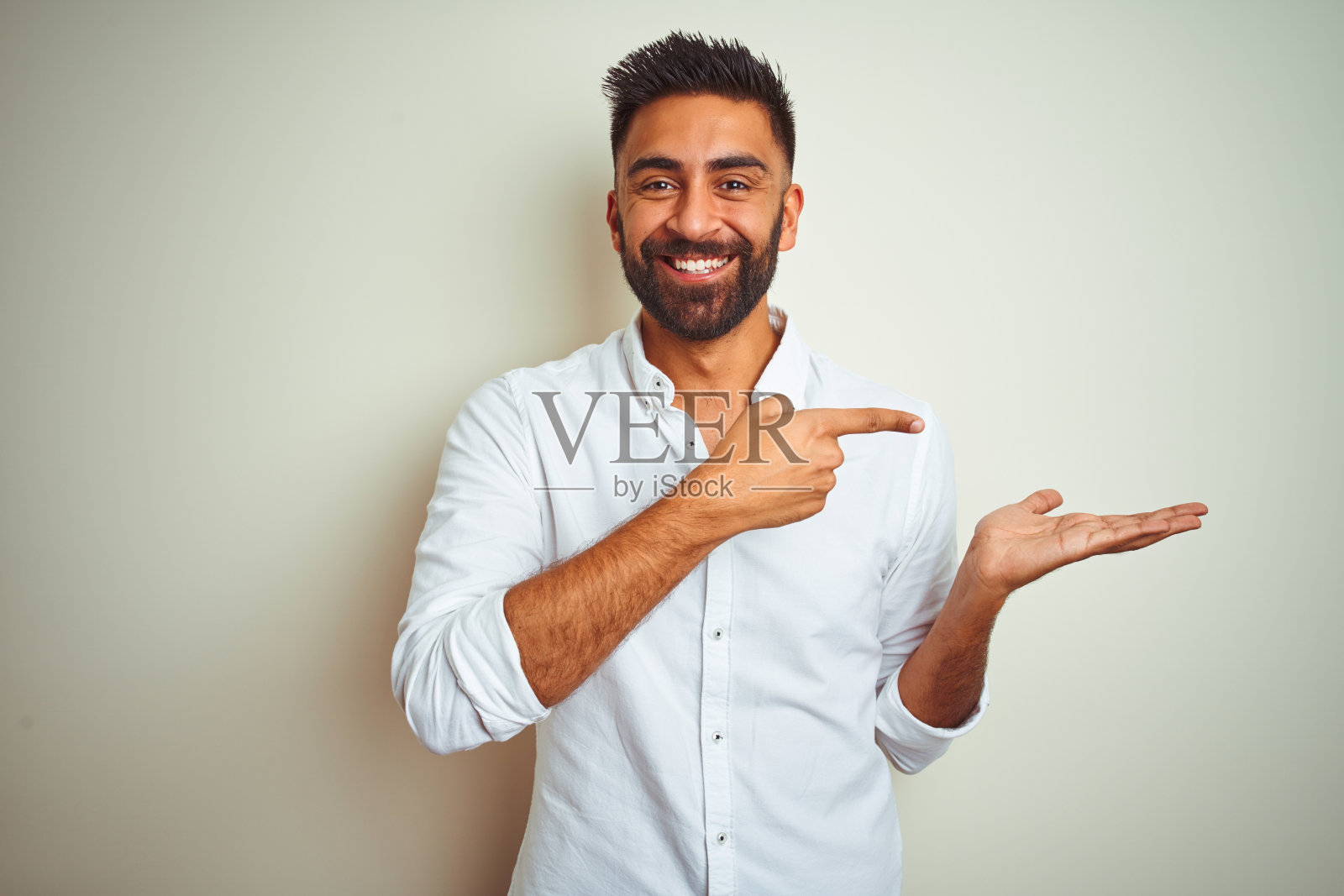 穿着优雅衬衫的年轻印度男子，站在白色背景前，惊讶地笑着看着镜头，用手展示并用手指指着。照片摄影图片