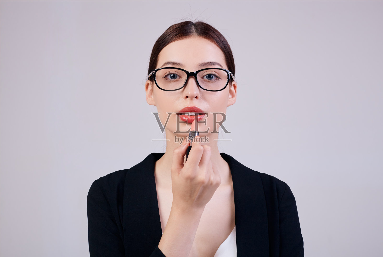 这位看起来和蔼冷静的商务女性右手涂着粉红色的口红，身穿黑色夹克，白色t恤，戴着电脑眼镜，站在灰色的后背上。忙碌的工人。副本的空间。照片摄影图片
