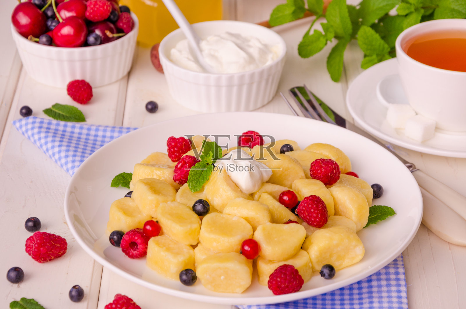 在白色的木质背景上，用新鲜的浆果、薄荷叶、酸奶油和蜂蜜做成的松软奶酪饺子。俄罗斯和乌克兰的传统早餐。水平,密切起来。照片摄影图片