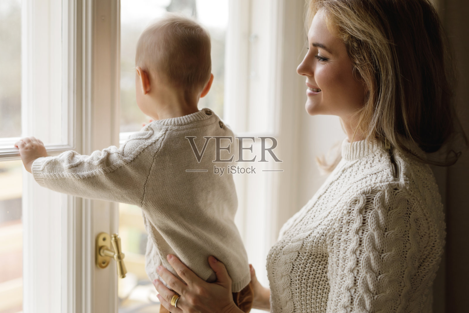 母亲和她穿着暖和毛衣的幼儿儿子在看窗外照片摄影图片
