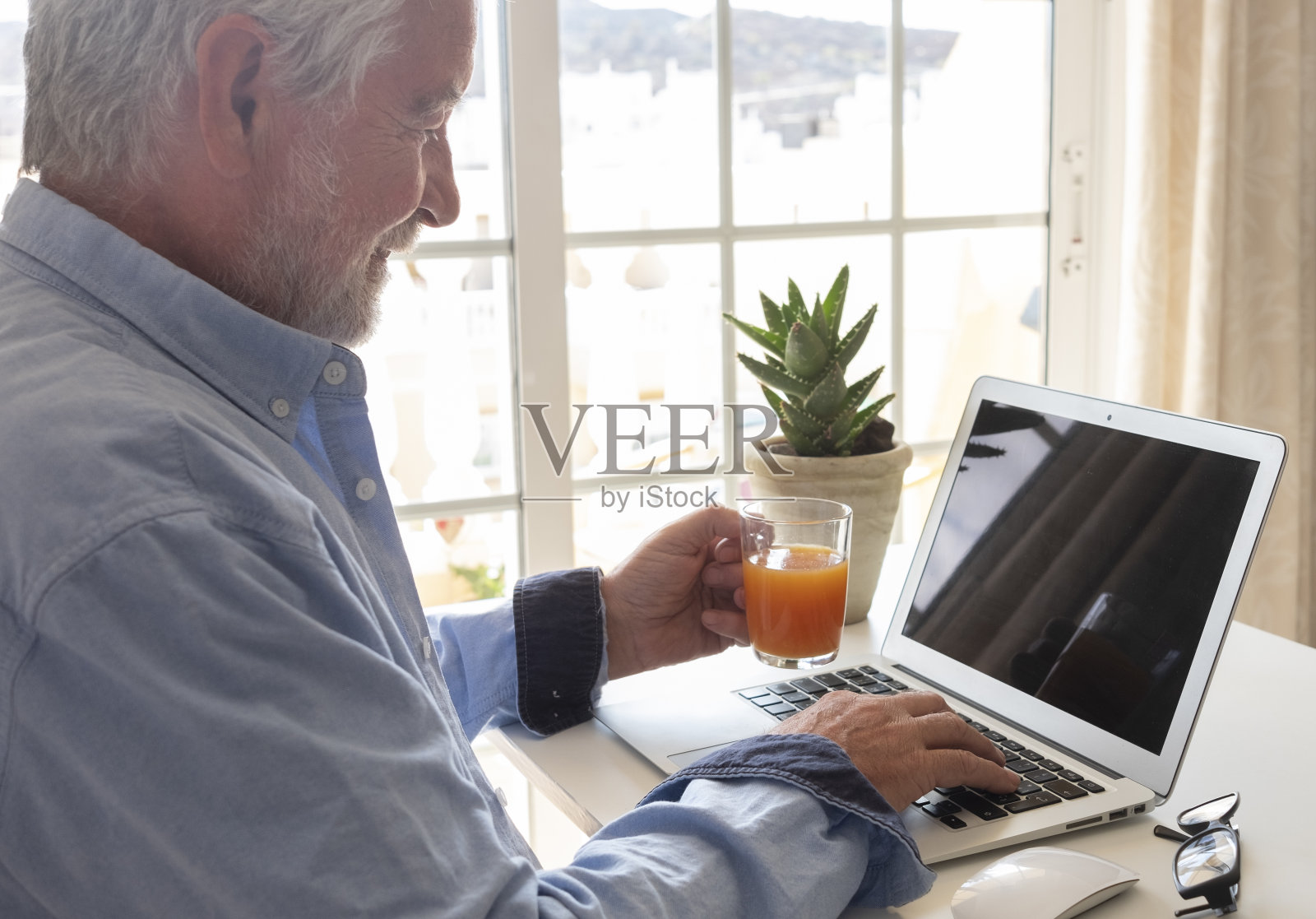 一个留着胡子和白发的老人正在用电脑——桌上的笔记本电脑和手机——从窗户透进来的亮光照片摄影图片