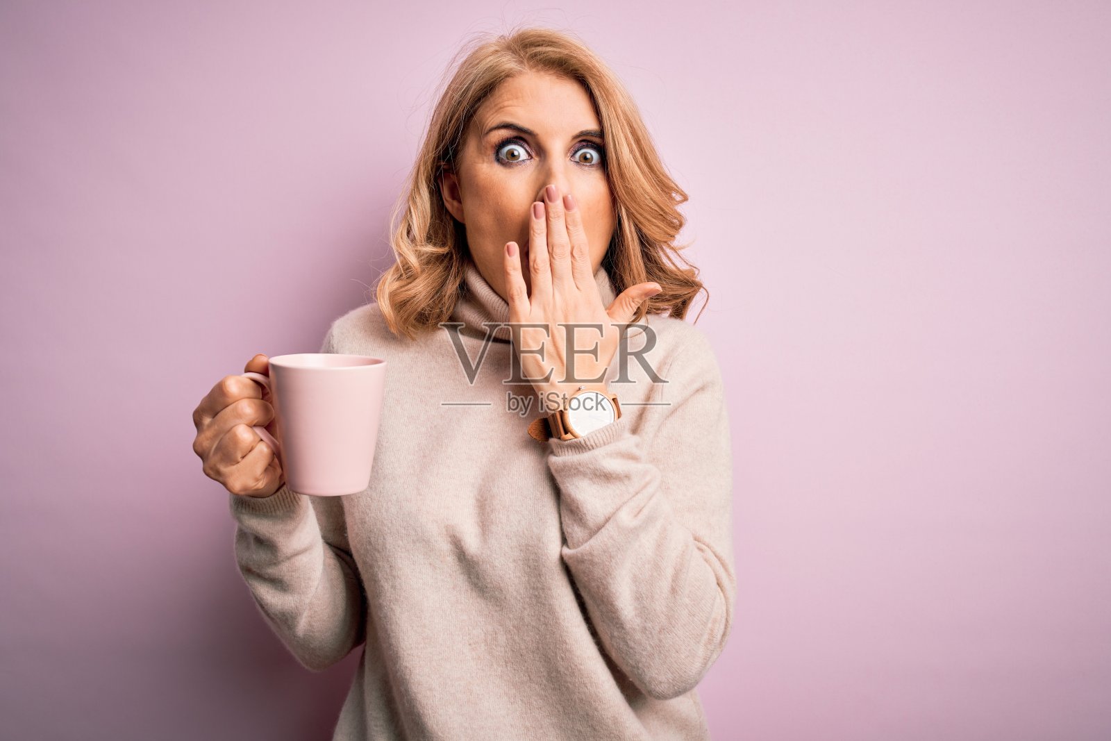 拿着粉色咖啡杯，因犯错而捂嘴震惊的中年金发美女照片摄影图片
