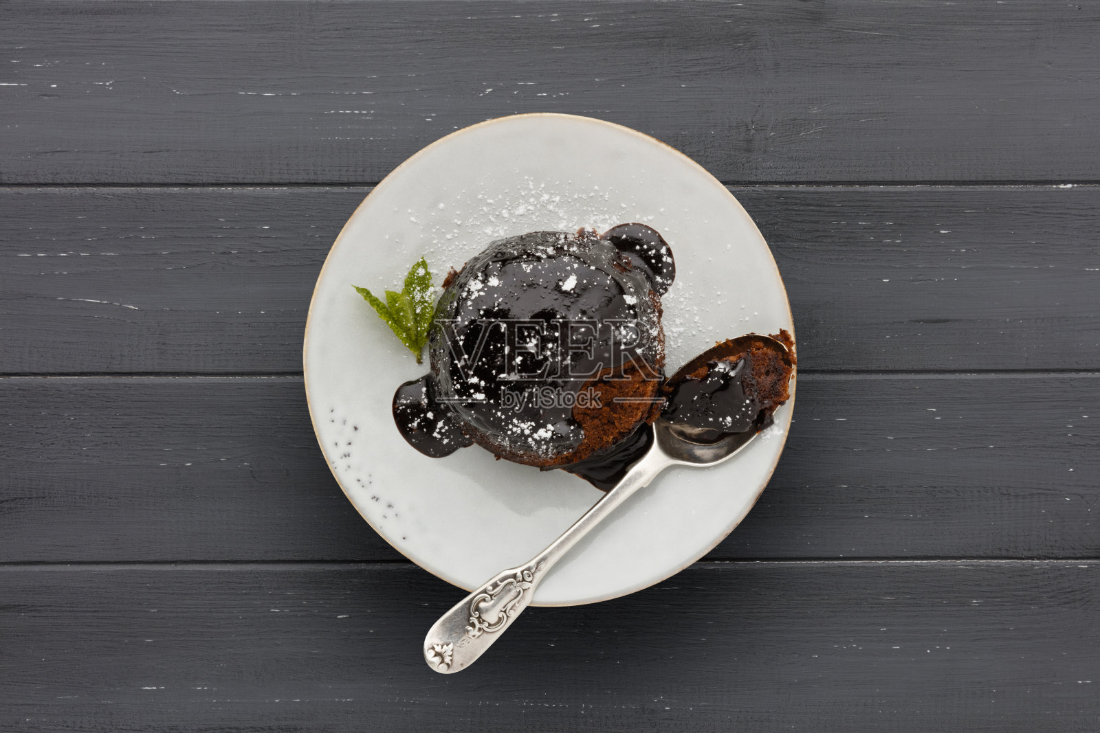 一个单独的巧克力海绵蛋糕，蘸着粘稠的巧克力酱，用勺子放在一个白色的盘子里，在有复制空间的深灰色木制表面上拍摄照片摄影图片