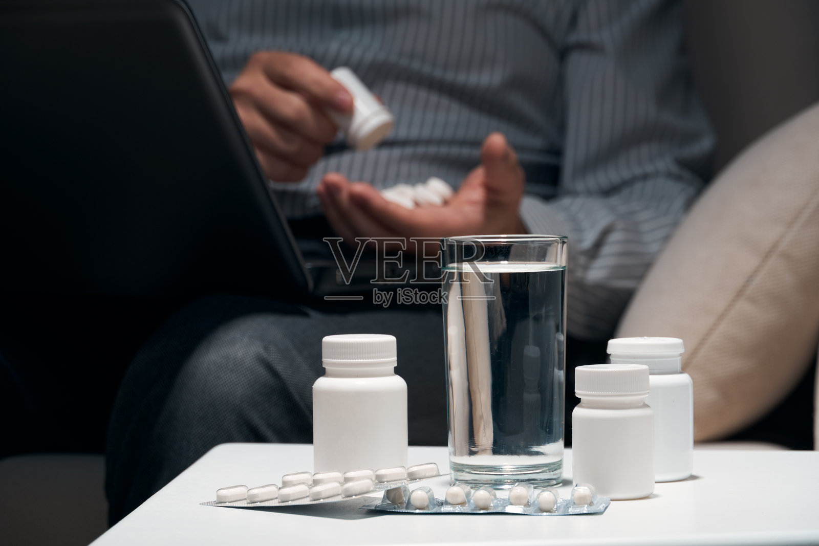 劳累过度的商人坐在沙发上吃药。药片和一杯水就在他身边。照片摄影图片