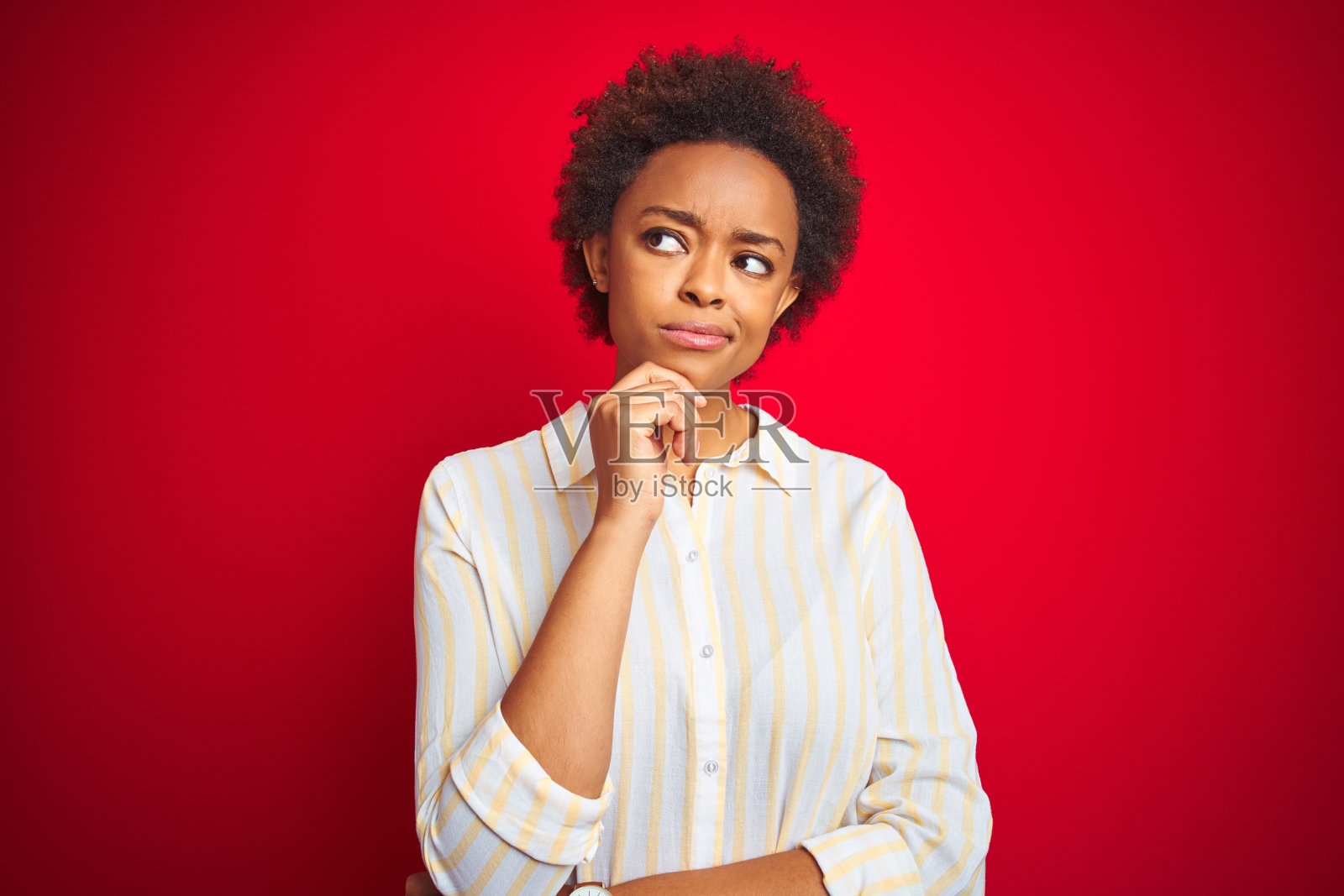 年轻漂亮的非裔美国女性，蓬松发型，红色背景，手托下巴思考问题，面露沉思，笑容满面，表情专注，疑问概念。照片摄影图片