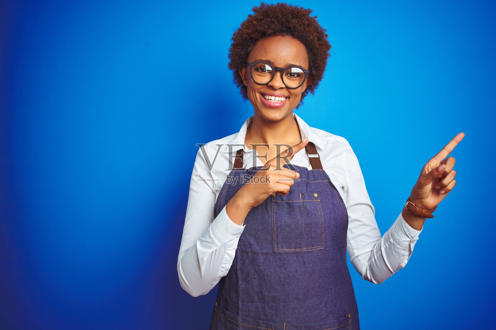 年轻的非裔美国女店主穿着工作围裙，在蓝色背景前微笑看着相机，用双手和手指指向侧面照片摄影图片