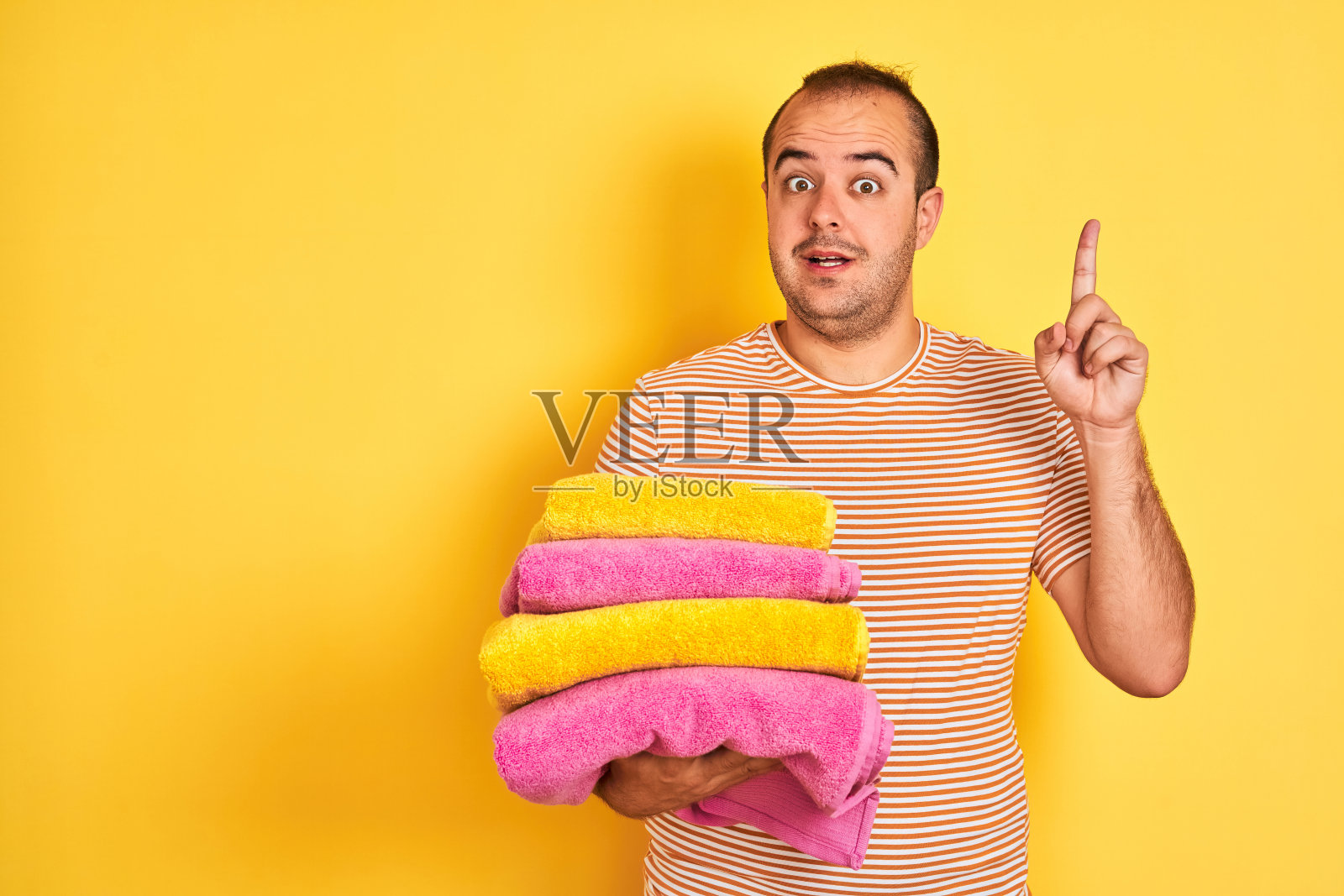 年轻的店主拿着折叠的毛巾站在孤立的黄色背景惊讶地提出一个想法或问题，用幸福的脸指着手指，第一个照片摄影图片