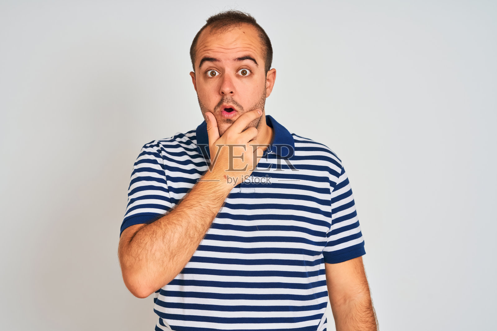 一个穿着休闲条纹马球衫的年轻人站在孤立的白色背景下，双手托着下巴，用难以置信、惊讶和惊讶的表情看着他照片摄影图片
