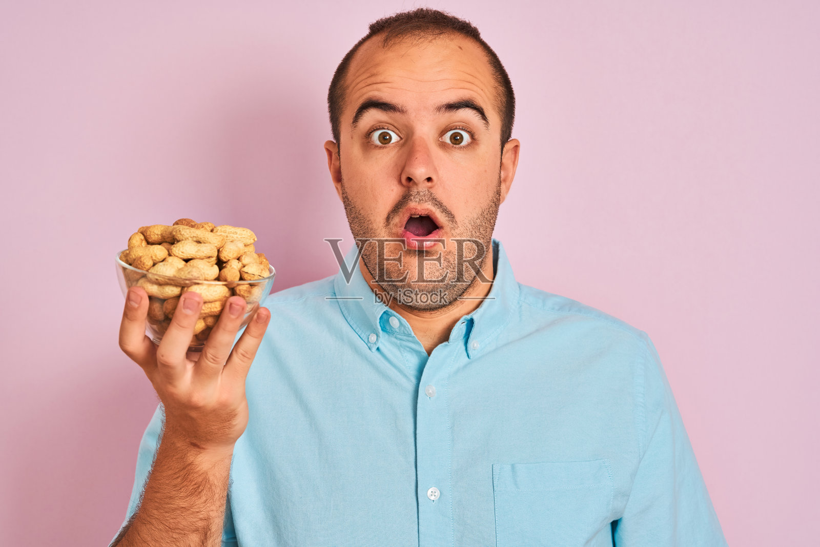 年轻人捧着碗里的花生站在孤立的粉红色背景下，吓得满脸惊讶，又怕又激动，带着恐惧的表情照片摄影图片