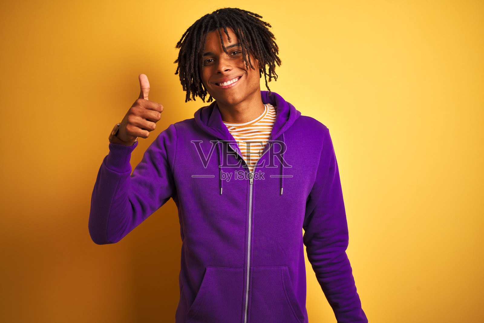非裔美国男人，头戴脏辫，身穿紫色运动衫，在紫色背景前，高兴地竖起大拇指。面带赞许的表情看着镜头，展示成功。照片摄影图片