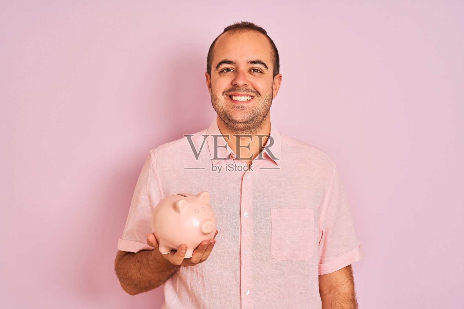 年轻男子拿着储蓄罐站在孤立的粉红色背景与快乐的脸站着，微笑着自信的微笑显示牙齿照片摄影图片