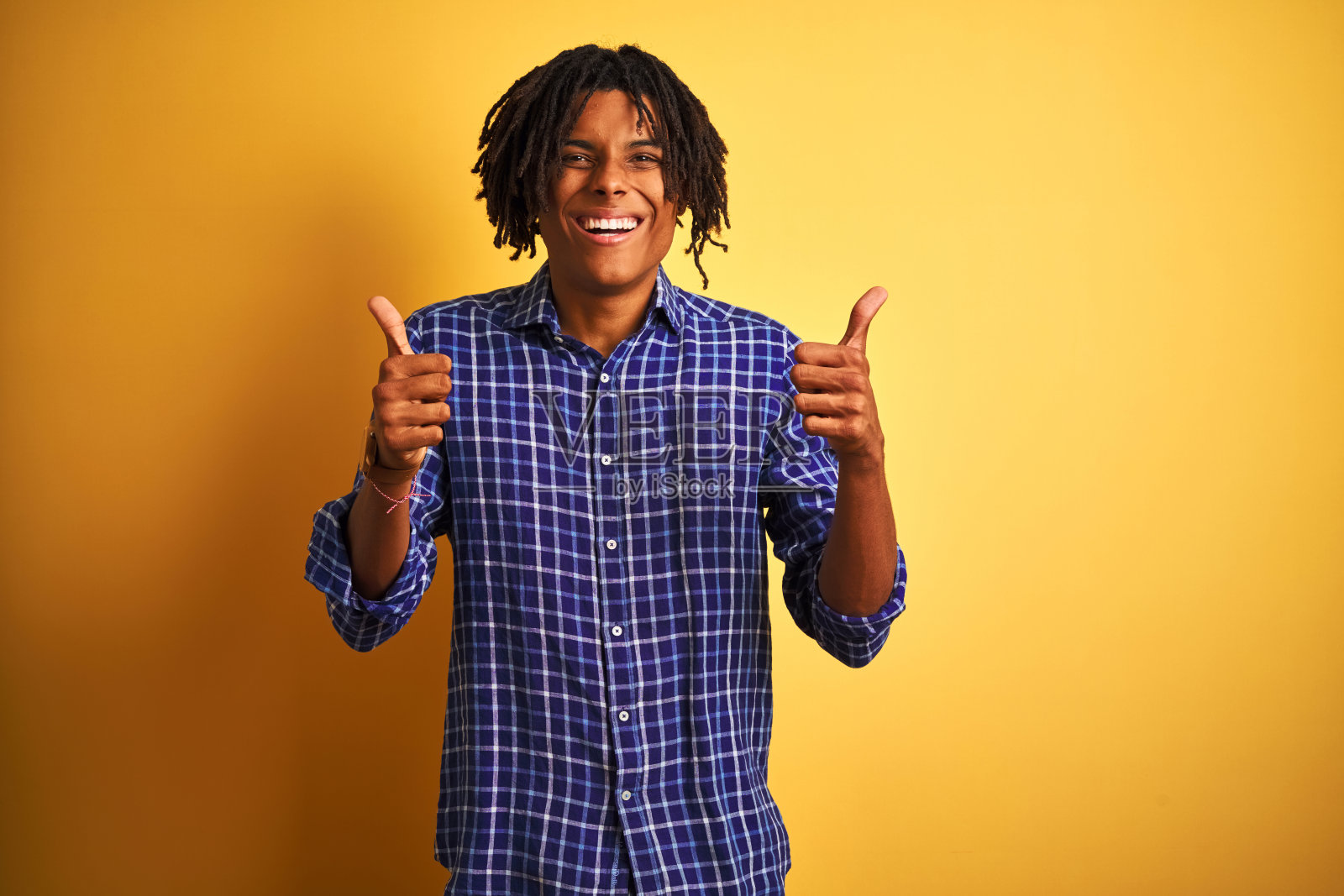 黑人男子，留着脏辫，穿着休闲衬衫，在黄色背景前竖起大拇指，面带微笑，表达快乐，庆祝成功照片摄影图片